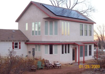 A two-story tan home addition with red trim and rooftop solar panels, overlooking a dirt yard with outdoor seating